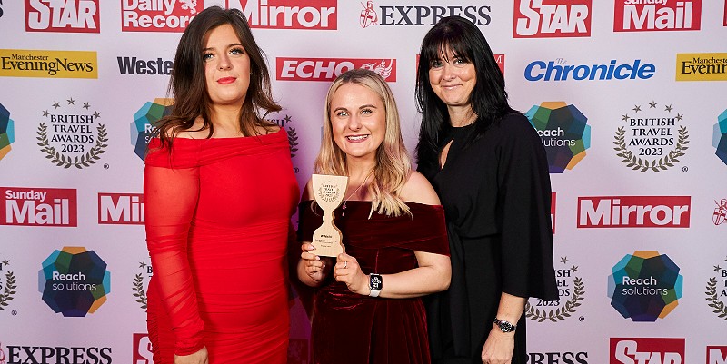 Three women from Blue Bay Travel pose with an award in front of advertising hordings