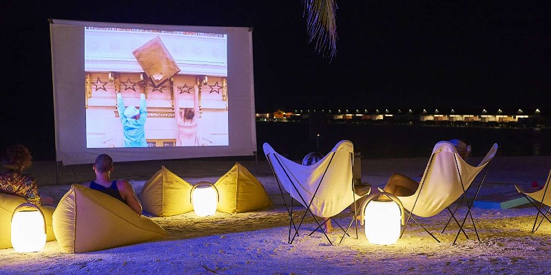 Two people watching a film on a projector screen at night on a Maldives beach
